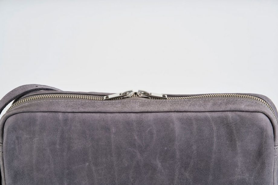 Zoom on a Product Picture view of a standing up blue vegetable tanned leather shoulder bag with a double silver zipper opening and an adjustable and detachable shoulder strap with a hook on one side and a buckle at the other side