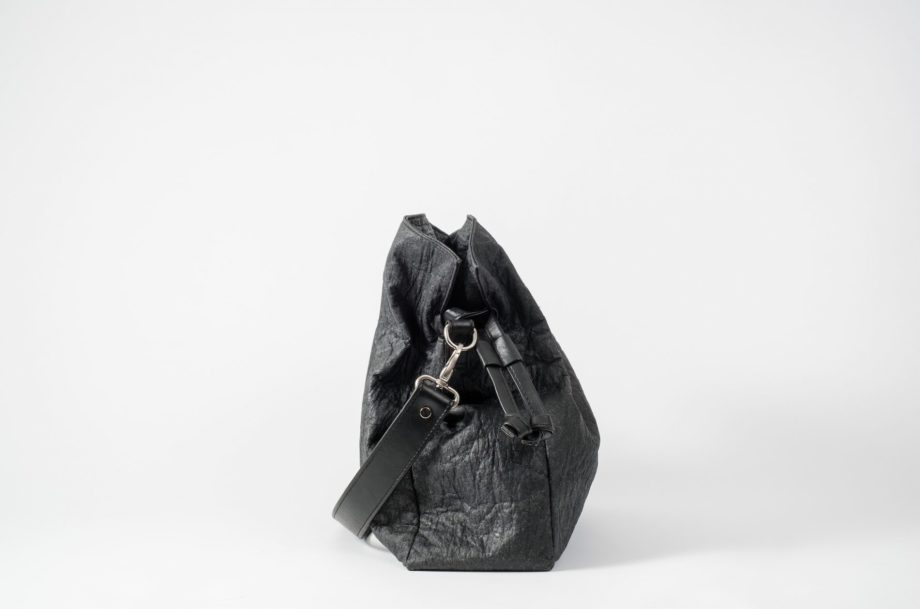 Side view on an closed black Pinatex Bucket-Tote bag with a detachable shoulder strap made of vegan leather dropped in front of the bag and a drawstring on the side of the bag