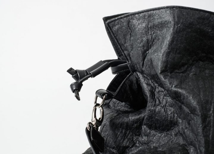Pinatex Totem Bag Zoom on the side top of a black Pinatex Bucket-Tote bag with a a drawstring with two knots and a hook with a shoulder strap made of vegan leather attached to a D-Ring