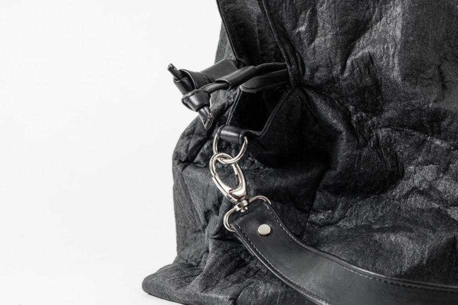 Zoom on the side of a black Pinatex Bucket-Tote bag with a a drawstring and a hook with a shoulder strap made of vegan leather attached to a D-Ring