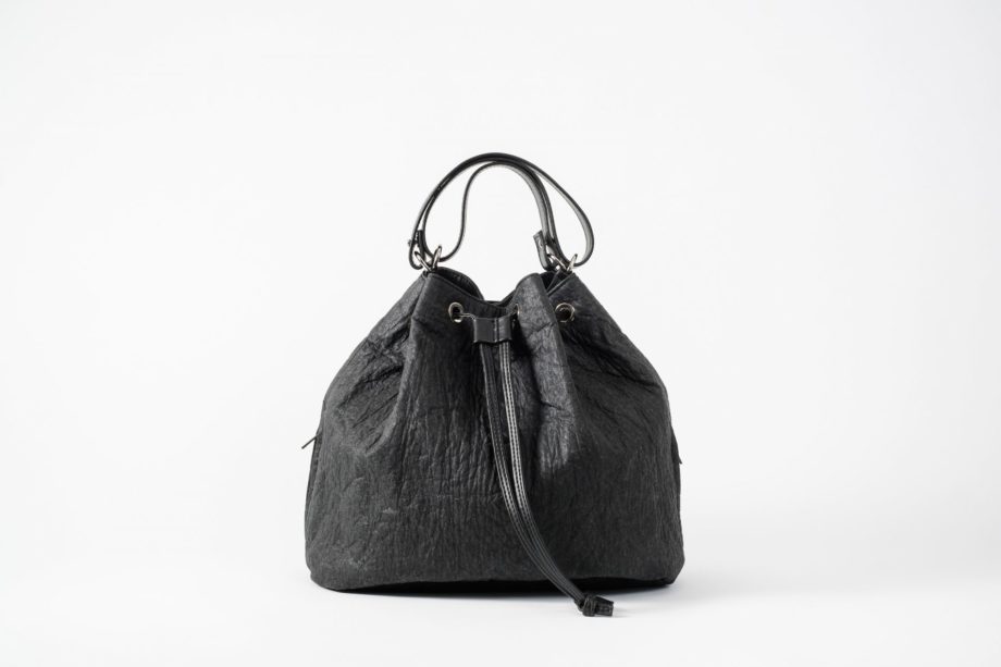 Front view on a standing up closed black Pinatex Bucket Bag with a zip pocket sewn in each side seam, a three times folded adjustable handle made of vegan leather and a drawstring knotted in the front