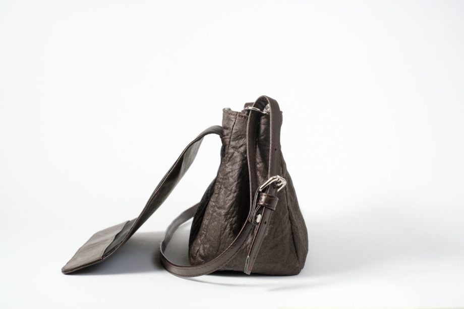 Side view of the open brown shoulder bag where as the body is made of pinatex and shows the side closed with a press button and the adjustable string and the back folded flap cover are made of vegan leather