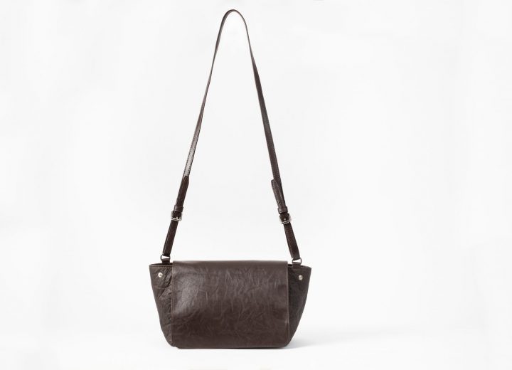 front view of an upstanding brown shoulderbag with a flap cover and adjustable string in vegan leather and the shape of the body of the bag is in Pinatex and can change volume by closing the press buttons on the sides