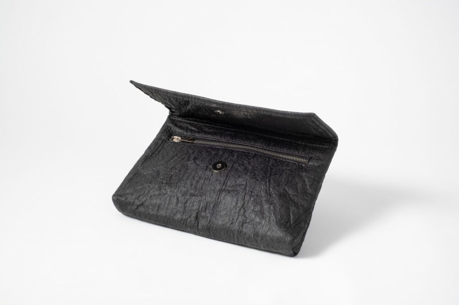 Front side view on an half standing charcoal colored Pinatex Clutch with the flap half open so that you can see the magnetic button and a zip opening